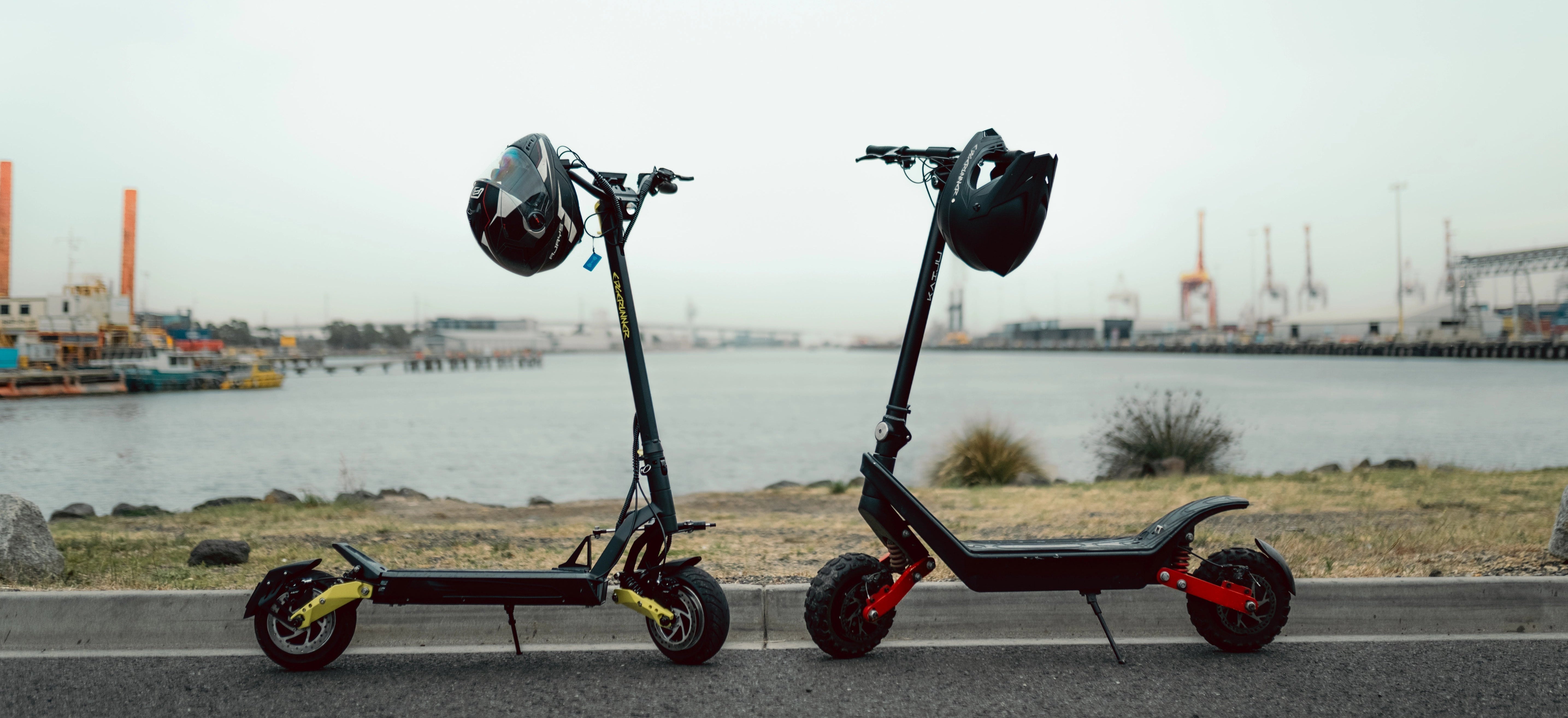 Two electric scooters on a road with a waterfront and industrial buildings in the background.